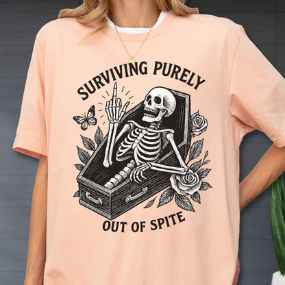 May include: A peach-colored t-shirt featuring a black graphic of a skeleton inside a coffin, giving the middle finger. The design includes roses and a butterfly, with the text "SURVIVING PURELY OUT OF SPITE" printed in a bold, arched black font.
