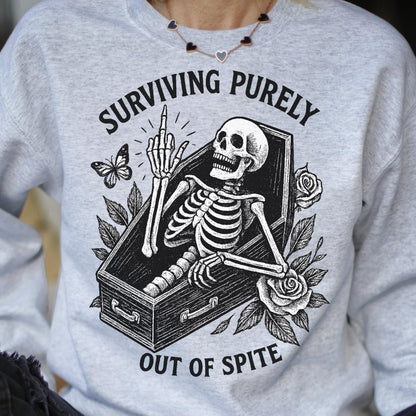 May include: A light gray sweatshirt featuring a black and white graphic of a skeleton in a coffin, giving the middle finger. The text reads "SURVIVING PURELY OUT OF SPITE" with a butterfly and roses.