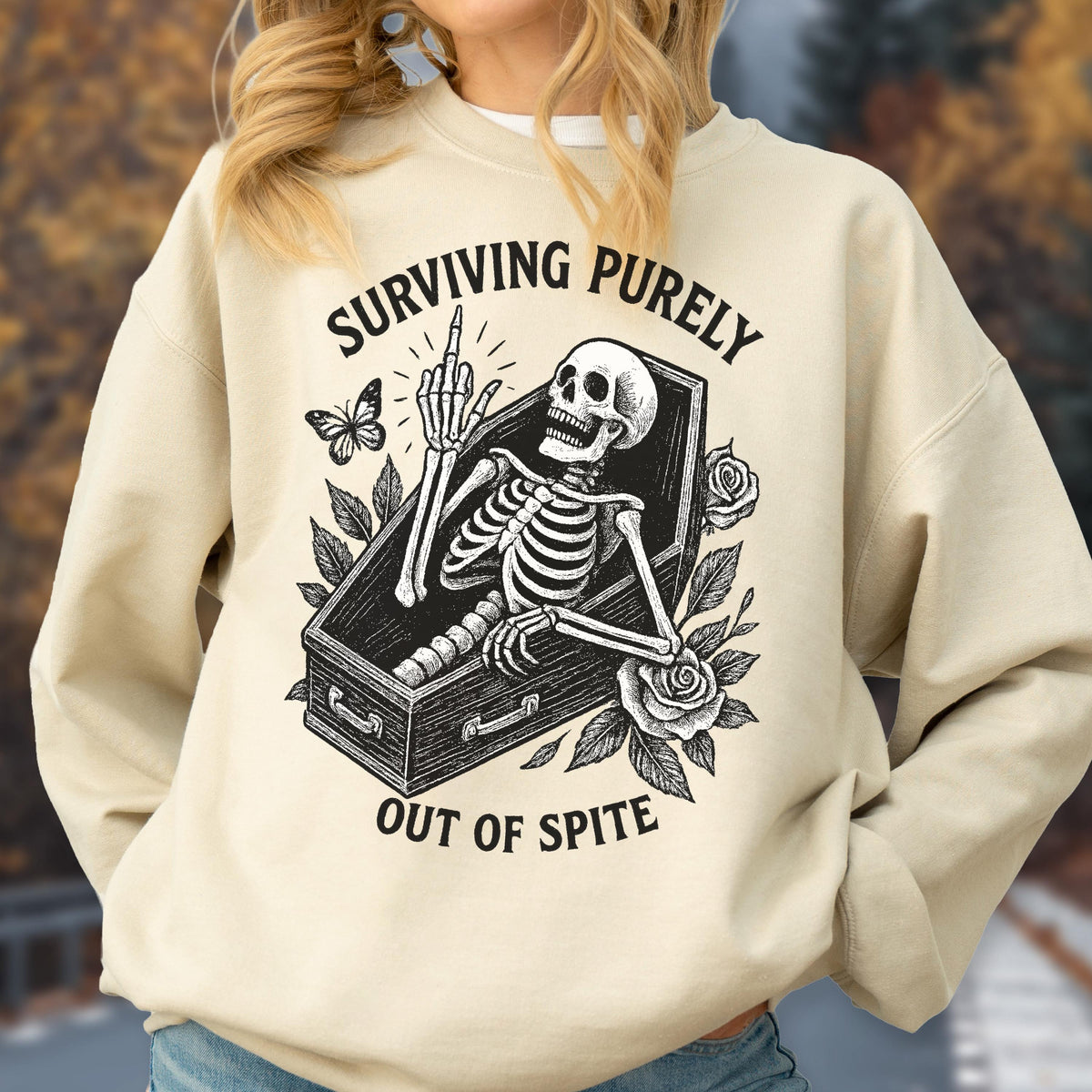 May include: Beige sweatshirt with a graphic of a skeleton in a coffin, giving the middle finger. The text reads "SURVIVING PURELY OUT OF SPITE". A butterfly and roses are also featured in the design.
