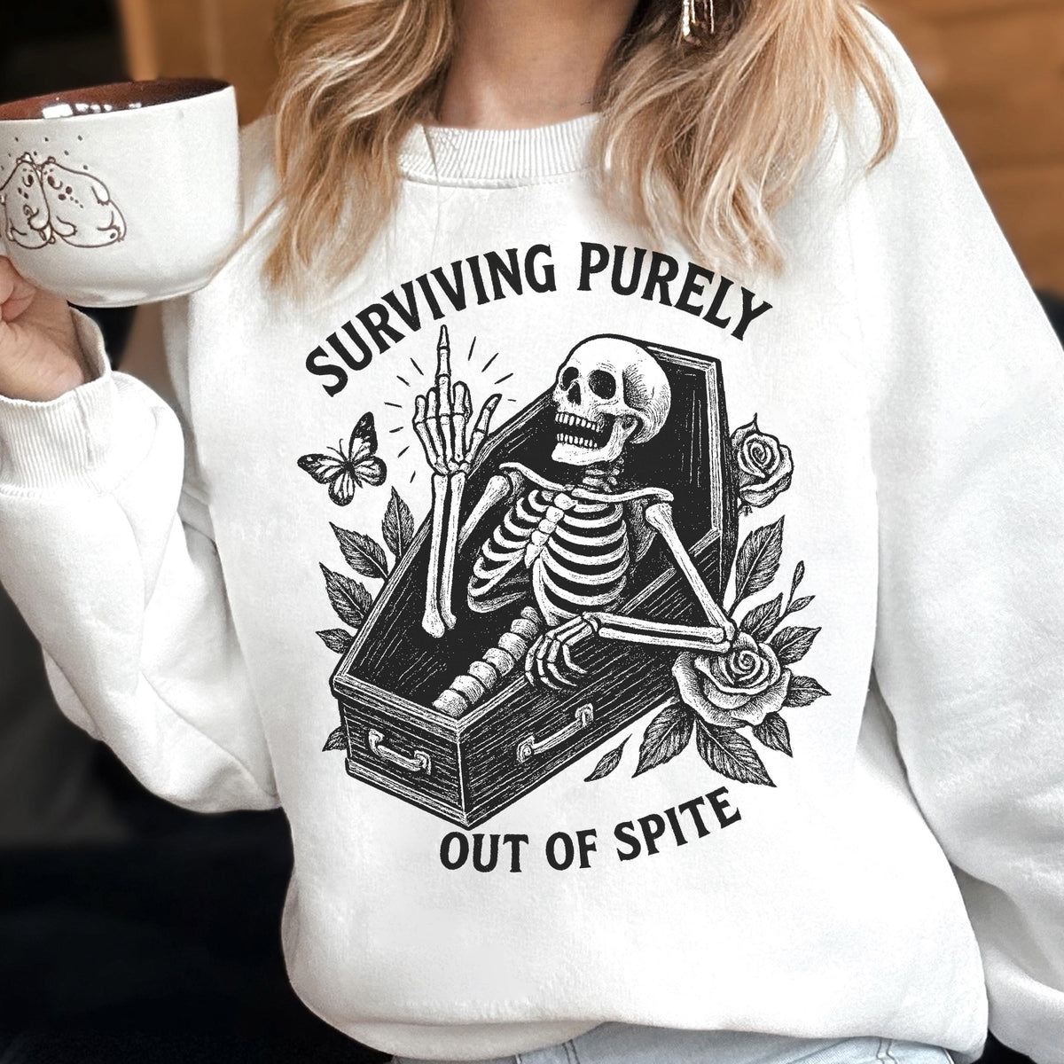 May include: White sweatshirt with a black and white graphic of a skeleton in a coffin, giving the middle finger. The text reads "SURVIVING PURELY OUT OF SPITE". A butterfly and roses are also in the design.