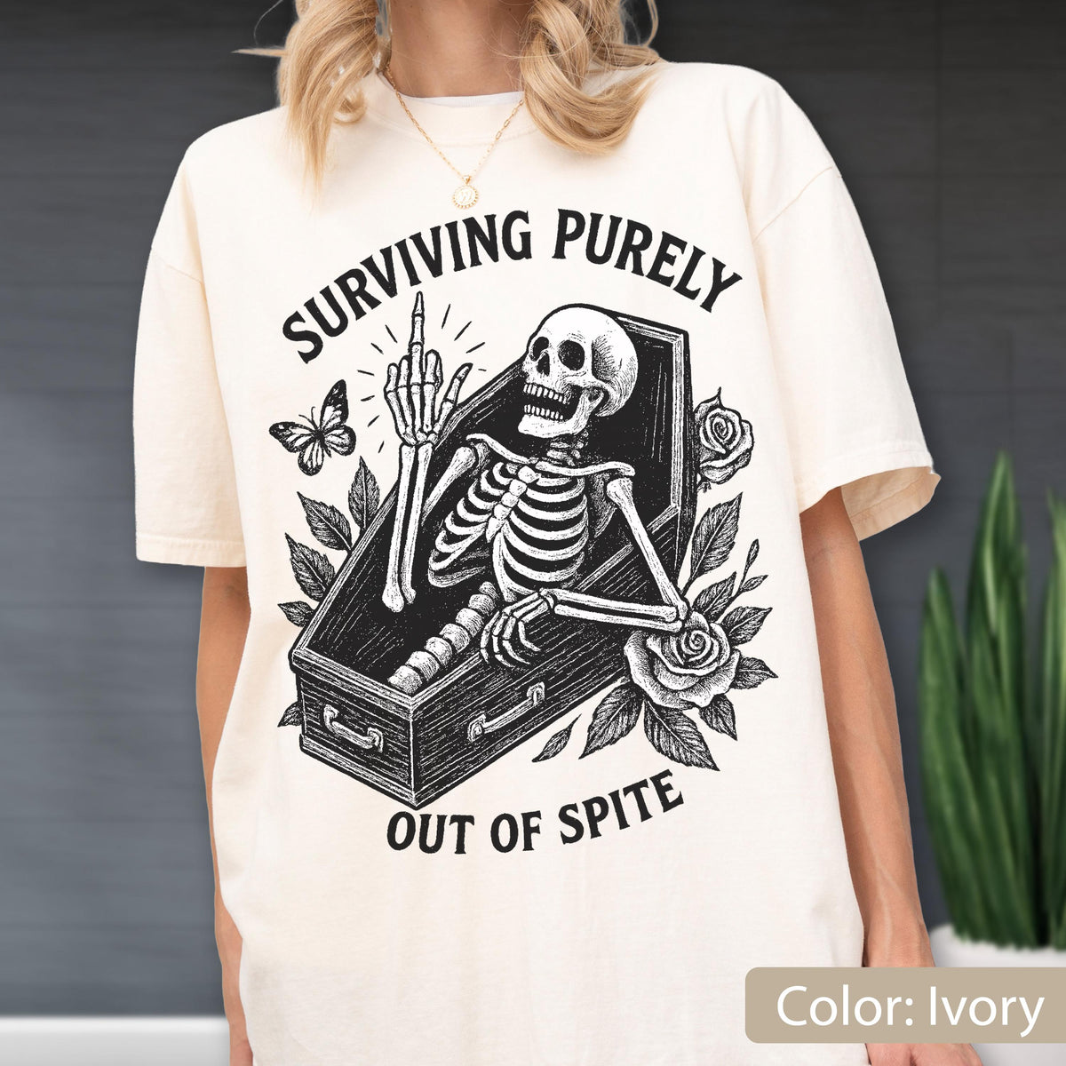 May include: An ivory t-shirt featuring a black graphic print of a skeleton inside a coffin, giving the middle finger. The design includes roses, a butterfly, and the text "SURVIVING PURELY OUT OF SPITE" arched above and below the central illustration.