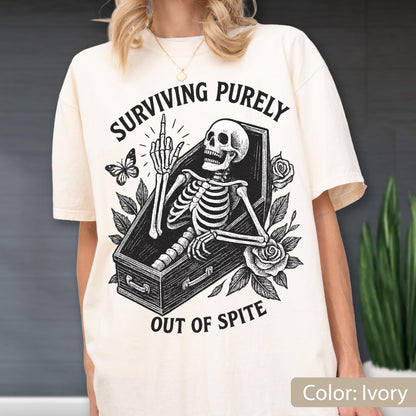 May include: An ivory t-shirt featuring a black graphic print of a skeleton inside a coffin, giving the middle finger. The design includes roses, a butterfly, and the text "SURVIVING PURELY OUT OF SPITE" arched above and below the central illustration.