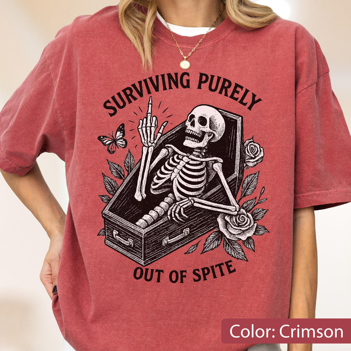May include: A crimson short-sleeve t-shirt featuring a black graphic of a skeleton inside a coffin, surrounded by roses and a butterfly. The skeleton is raising its middle finger. The text reads, "SURVIVING PURELY OUT OF SPITE" in black lettering.