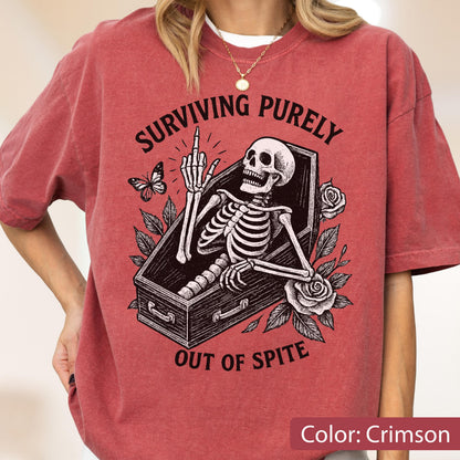 May include: A crimson short-sleeve t-shirt featuring a black graphic of a skeleton inside a coffin, surrounded by roses and a butterfly. The skeleton is raising its middle finger. The text reads, "SURVIVING PURELY OUT OF SPITE" in black lettering.