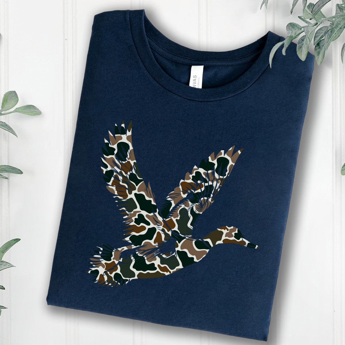 May include: A navy blue short-sleeved t-shirt featuring a graphic of a flying duck. The duck silhouette is filled with a camouflage pattern consisting of brown, dark green, and black organic shapes. The shirt has a crew neckline and a white label.