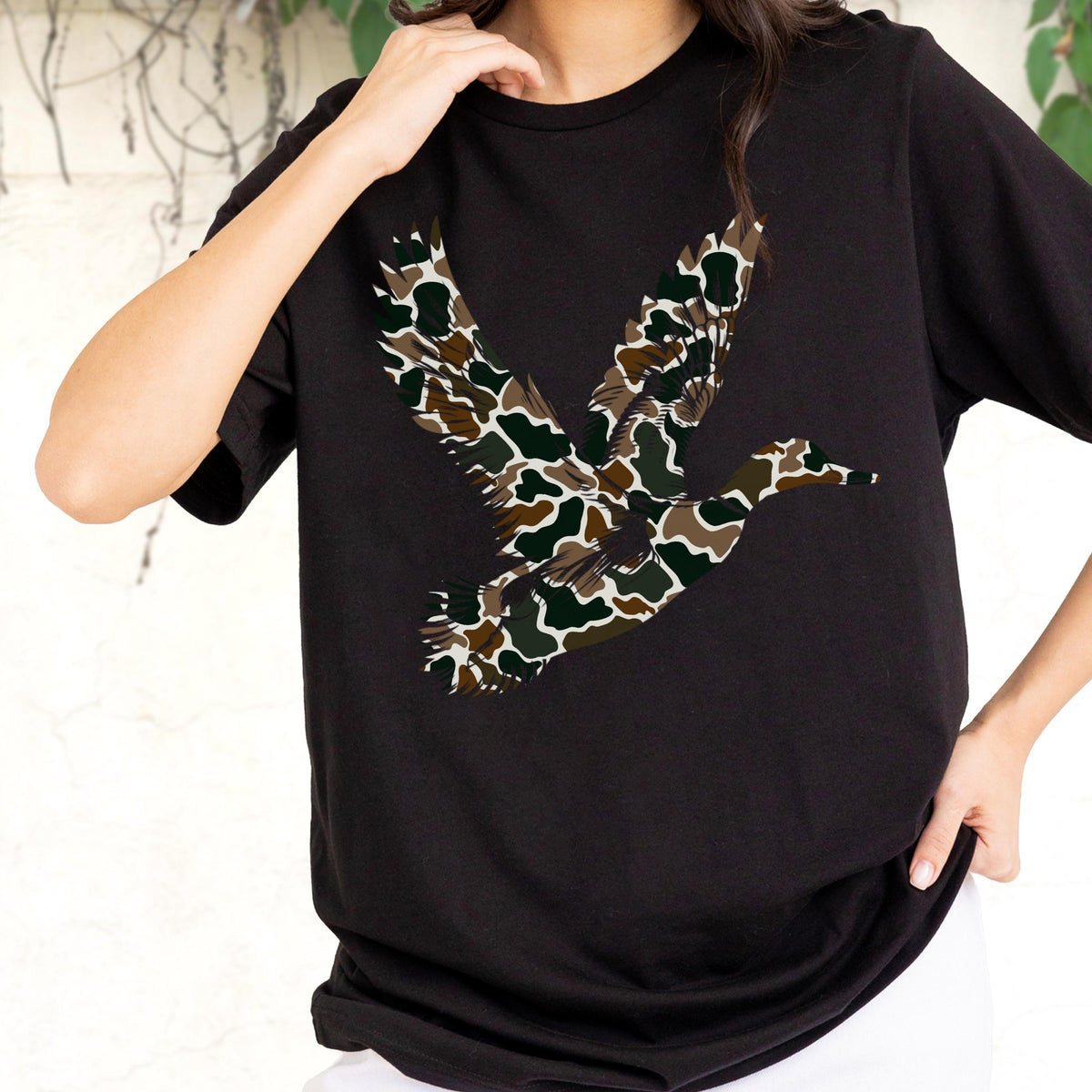 May include: A black short-sleeved t-shirt featuring a large graphic of a flying duck in the center. The duck silhouette is filled with a camouflage pattern consisting of dark green, brown, and tan organic shapes on a black background.