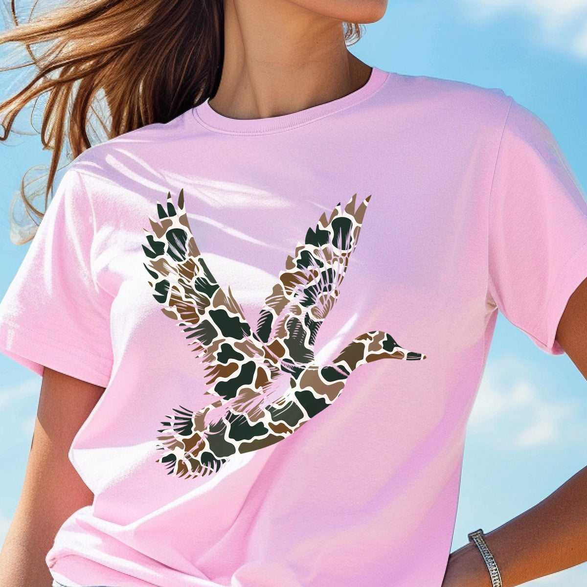 May include: A light pink short-sleeve t-shirt featuring a graphic of a flying duck. The duck is filled with a camouflage pattern consisting of brown, tan, dark green, and black organic shapes. The shirt has a crew neckline and a standard fit.
