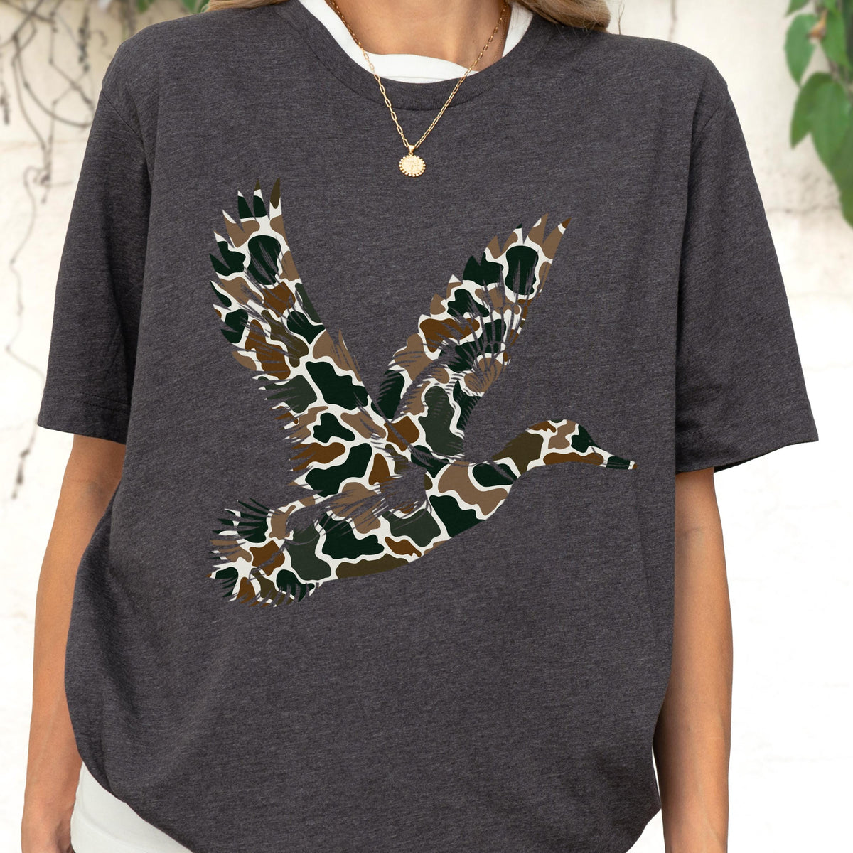 May include: A charcoal gray short-sleeved t-shirt featuring a large graphic of a flying duck. The duck silhouette is filled with a camouflage pattern consisting of brown, dark green, and cream-colored organic shapes across the entire body.