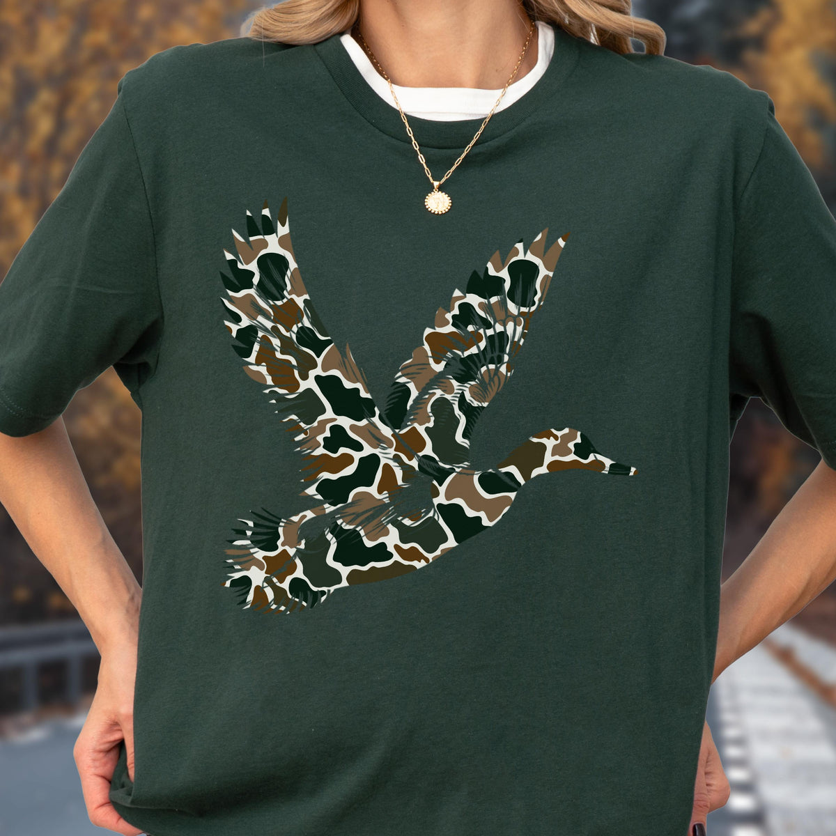 May include: A dark forest green short-sleeve t-shirt featuring a large graphic of a flying duck. The duck silhouette is filled with a camouflage pattern consisting of brown, tan, black, and dark green organic shapes across its wings and body.