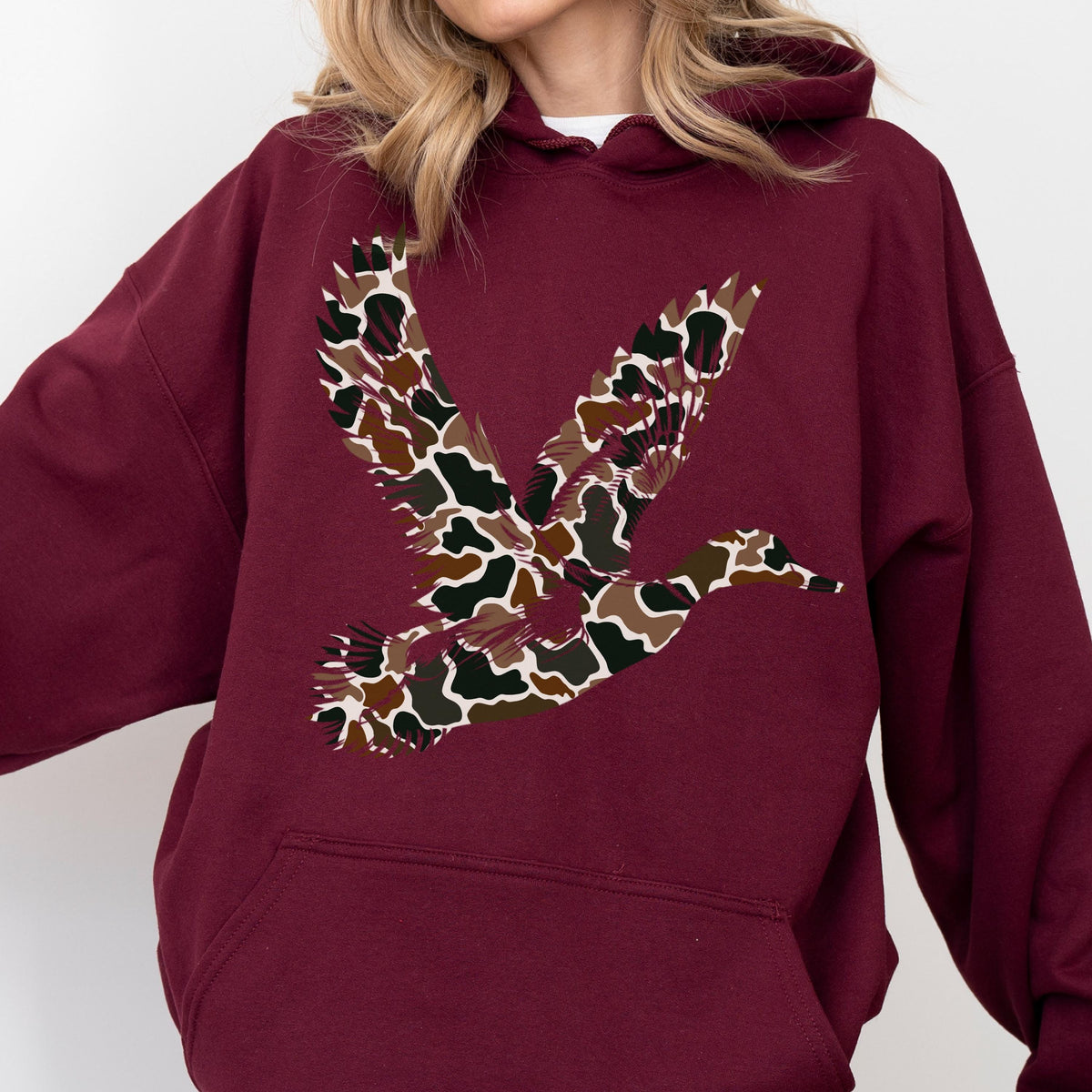 May include: A maroon hooded sweatshirt featuring a large graphic of a flying duck on the chest. The duck silhouette is filled with a multi-colored camouflage pattern consisting of brown, tan, black, and dark green organic shapes and spots.