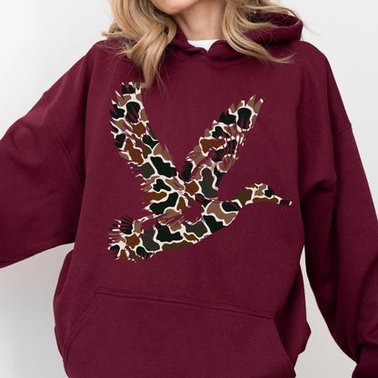 May include: A maroon hooded sweatshirt featuring a large graphic of a flying duck on the chest. The duck silhouette is filled with a multi-colored camouflage pattern consisting of brown, tan, black, and dark green organic shapes and spots.