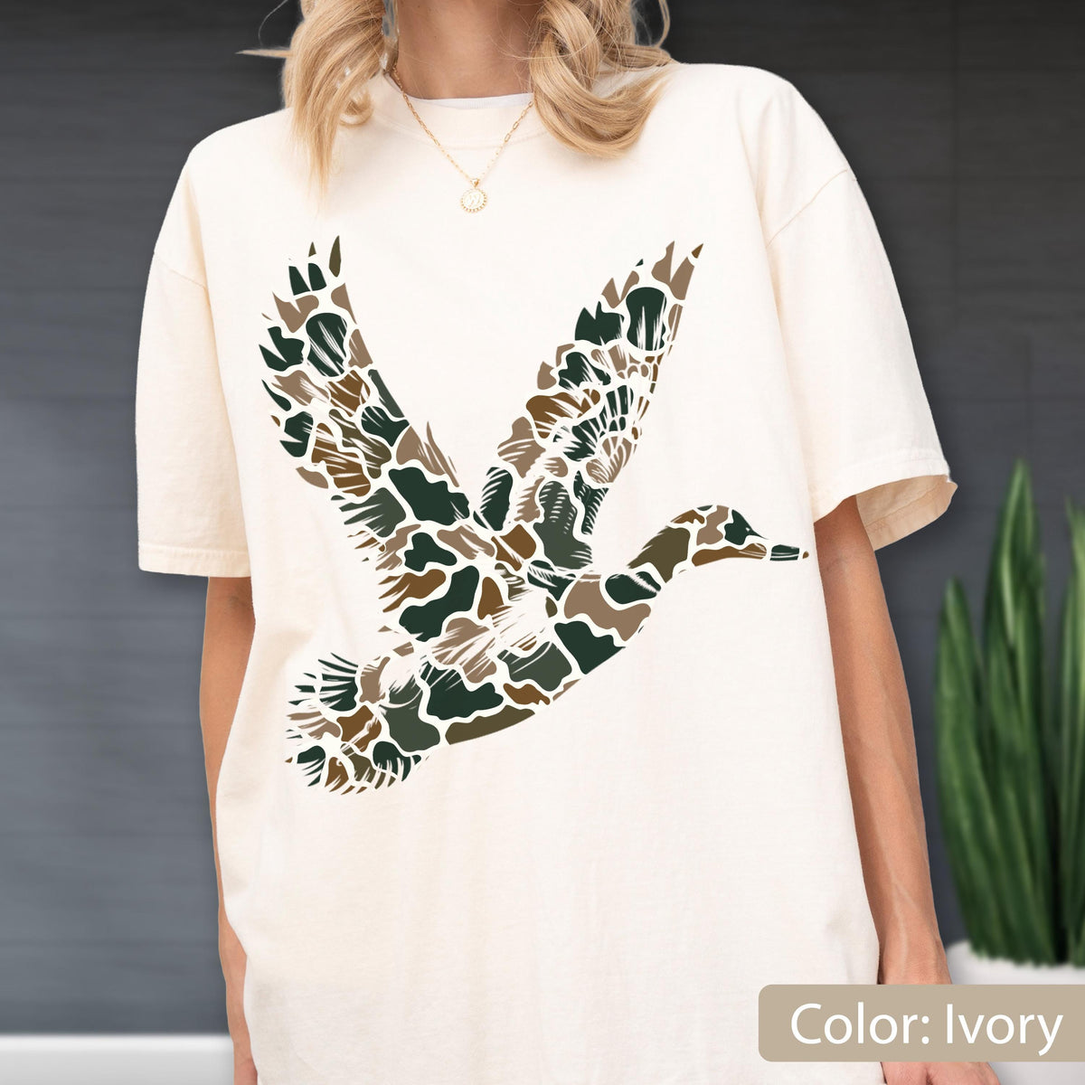 May include: An ivory short-sleeve t-shirt featuring a large graphic of a flying duck on the front. The duck silhouette is filled with a camouflage pattern consisting of dark green, brown, and tan organic shapes against the light fabric background.