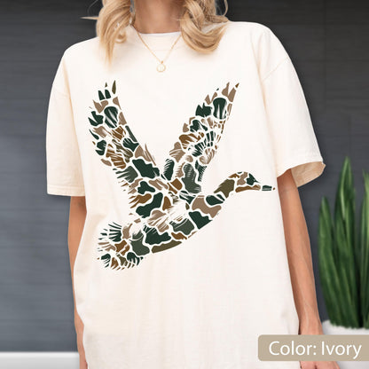 May include: An ivory short-sleeve t-shirt featuring a large graphic of a flying duck on the front. The duck silhouette is filled with a camouflage pattern consisting of dark green, brown, and tan organic shapes against the light fabric background.