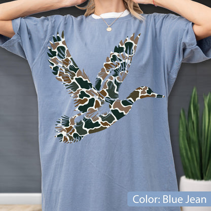 May include: A blue jean colored short-sleeve t-shirt featuring a large graphic of a flying duck. The duck silhouette is filled with a camouflage pattern consisting of dark green, brown, and white organic shapes across the chest area.
