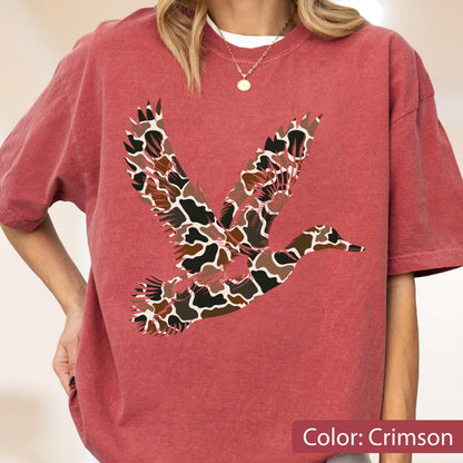 May include: A crimson short-sleeve t-shirt featuring a large graphic of a flying duck. The duck silhouette is filled with a brown, black, and white camouflage pattern. The shirt has a crew neckline and a relaxed fit, showcasing a bold animal design.