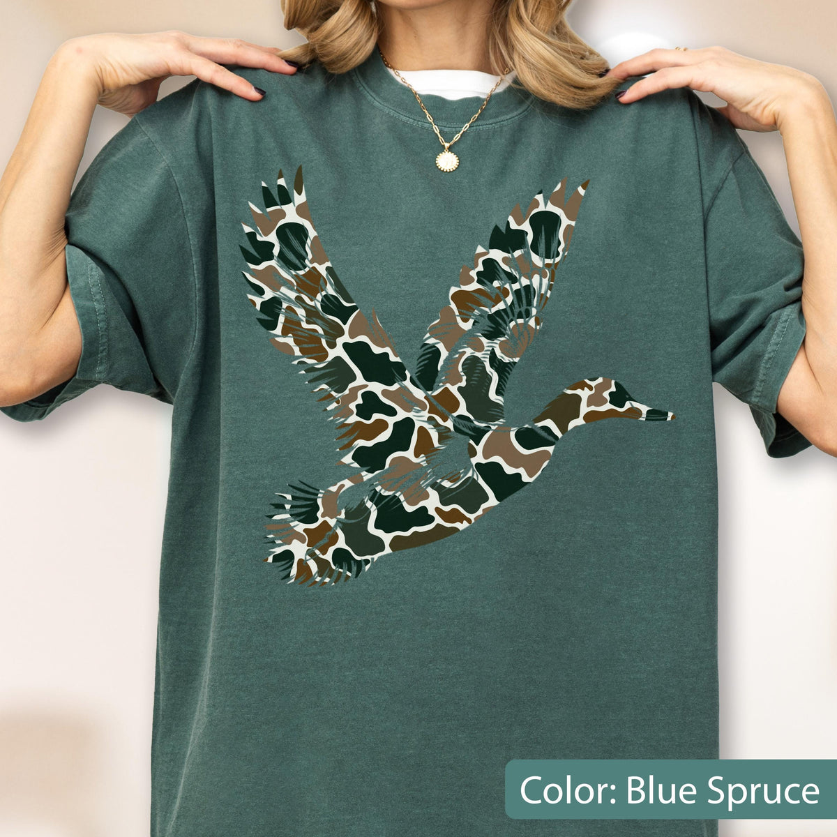 May include: A blue spruce short-sleeve t-shirt featuring a large graphic of a flying duck. The duck silhouette is filled with a camouflage pattern consisting of brown, dark green, and white organic shapes. The shirt has a crew neckline.
