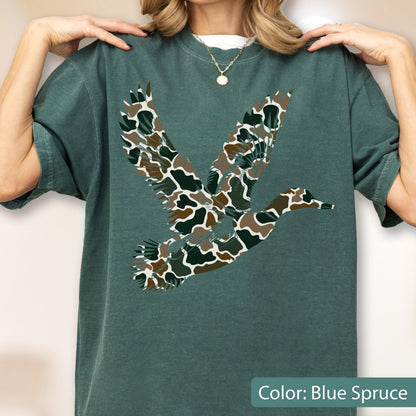 May include: A blue spruce short-sleeve t-shirt featuring a large graphic of a flying duck. The duck silhouette is filled with a camouflage pattern consisting of brown, dark green, and white organic shapes. The shirt has a crew neckline.