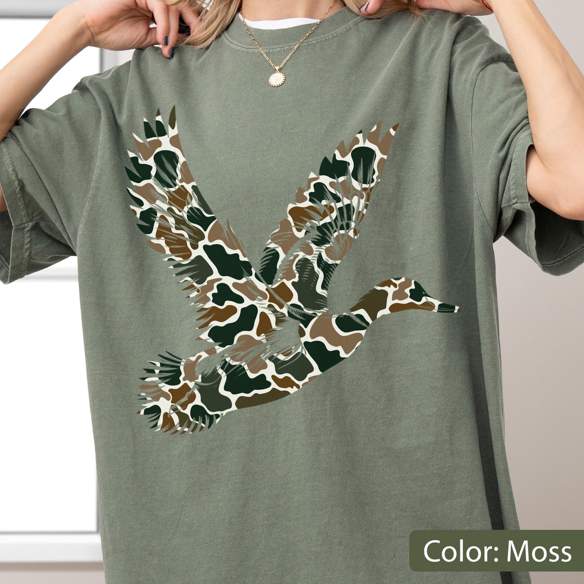 May include: A moss green short-sleeve t-shirt featuring a large graphic of a flying duck. The duck silhouette is filled with a camouflage pattern consisting of dark green, brown, and white organic shapes. The shirt has a crew neckline.