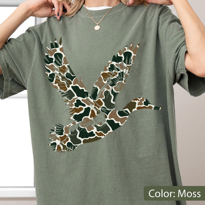 May include: A moss green short-sleeve t-shirt featuring a large graphic of a flying duck. The duck silhouette is filled with a camouflage pattern consisting of dark green, brown, and white organic shapes. The shirt has a crew neckline.