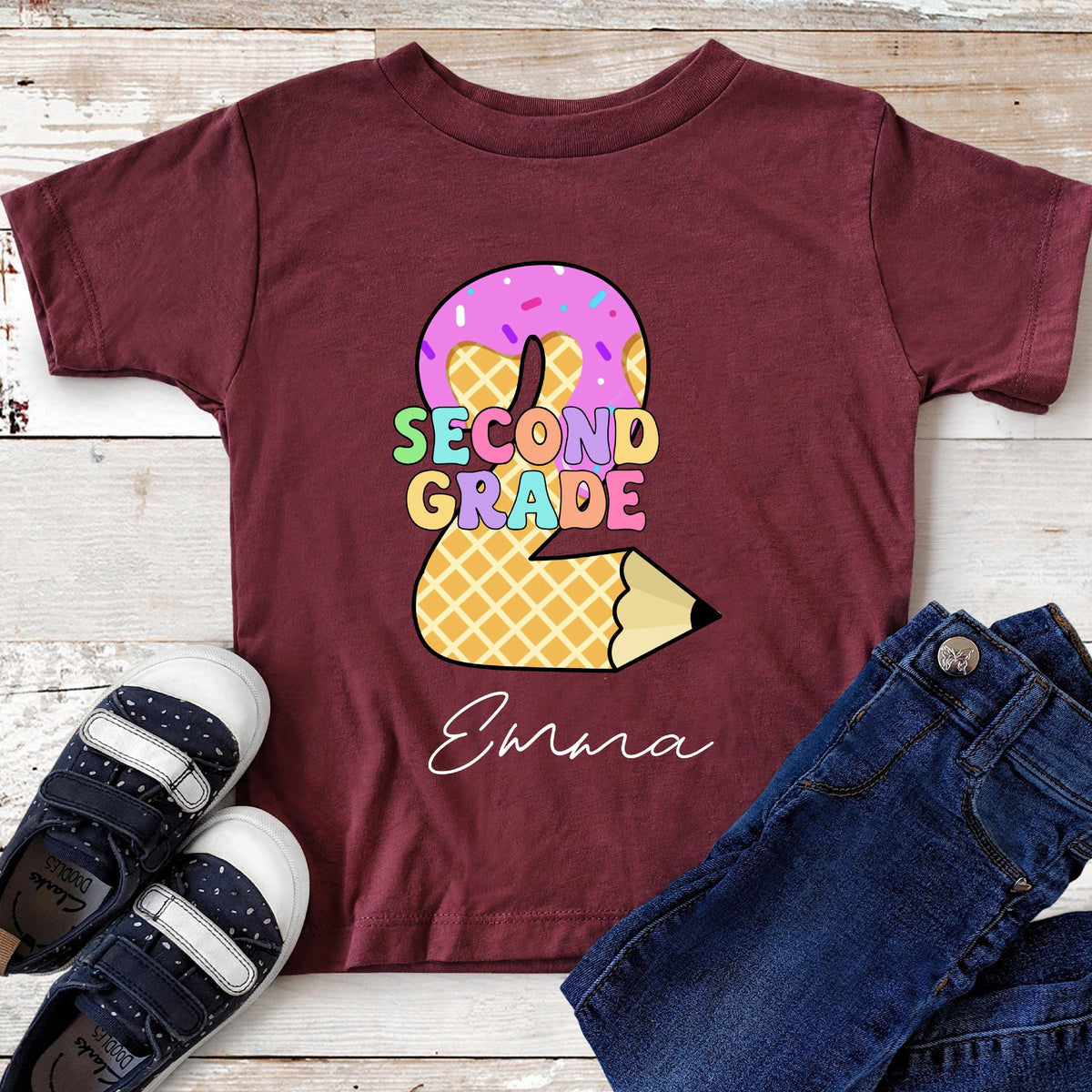 May include: Burgundy Second Grade t-shirt with a donut-shaped number 2 design. The donut is pink with sprinkles, and the number 2 is made of a waffle pattern. The words "SECOND GRADE" are in colorful letters, and the name "Emara" is written below.