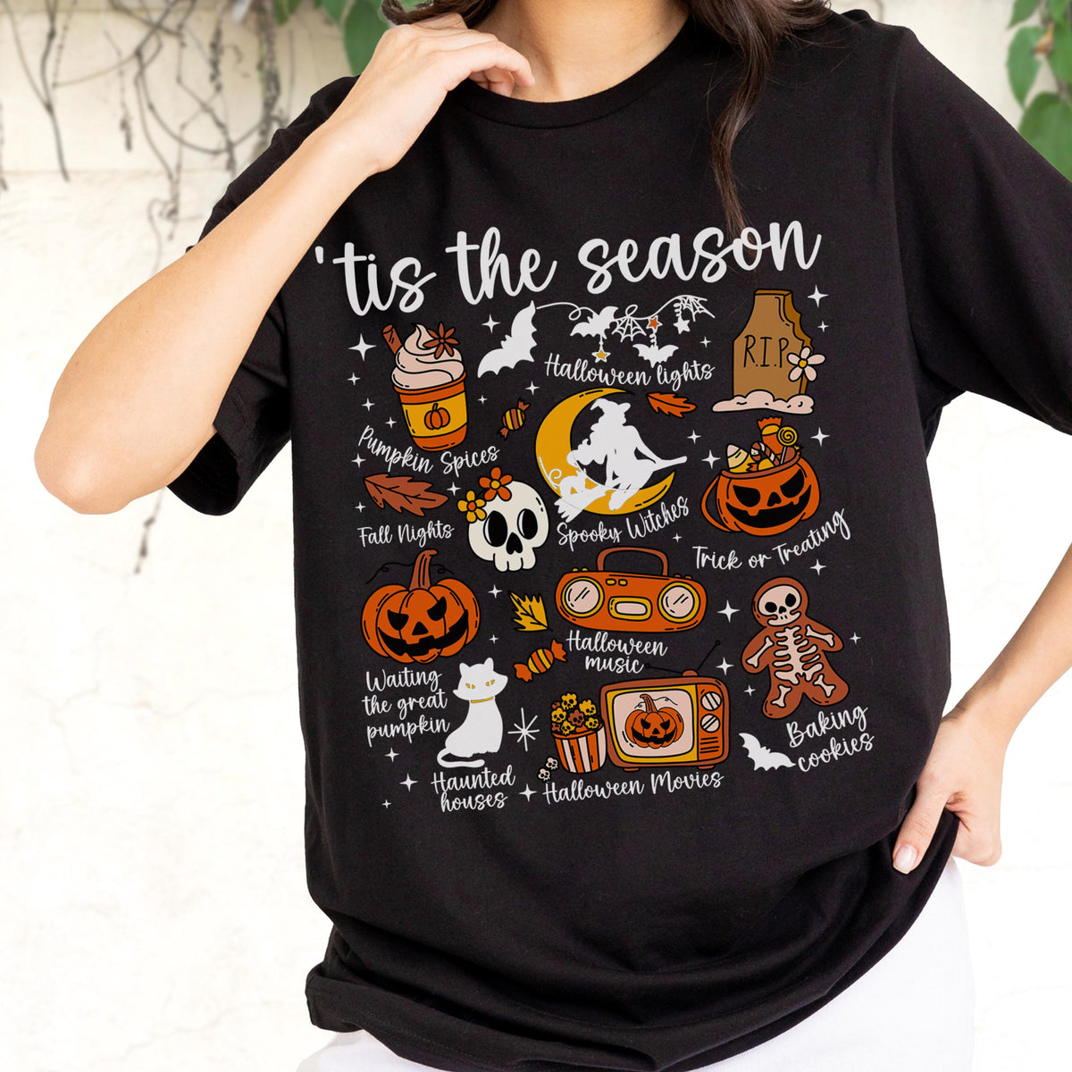 May include: Black t-shirt with a Halloween-themed graphic. The design includes pumpkins, bats, a skull, and the phrase "'tis the season." Other elements include a witch, a radio, and the words "Pumpkin Spices" and "Trick or Treating."