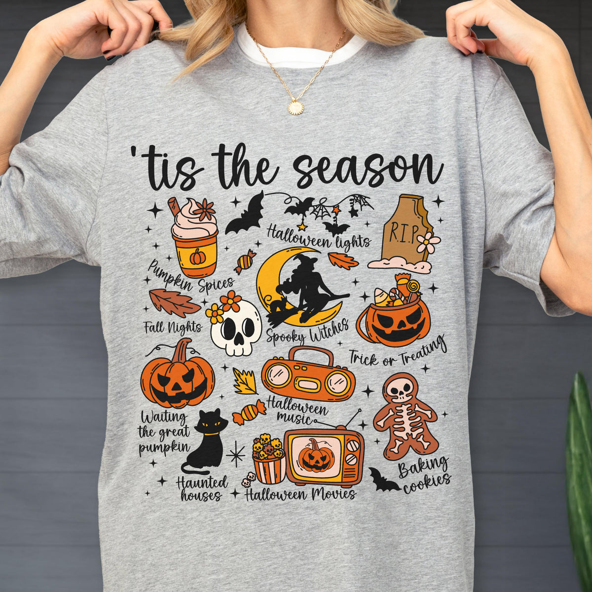 May include: A gray t-shirt with a Halloween-themed graphic. The design includes a variety of spooky elements such as pumpkins, bats, a skull, and the phrase "'tis the season". Additional text includes "Pumpkin Spices", "Spooky Witches", and "Trick or Treating".