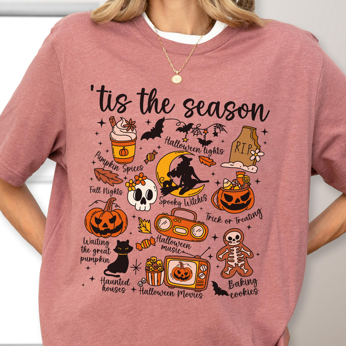 May include: Dusty rose t-shirt with a Halloween-themed graphic. The design includes the phrase "'tis the season" and various illustrations such as pumpkins, bats, a skull, and a witch on a crescent moon. The shirt is a crew neck.
