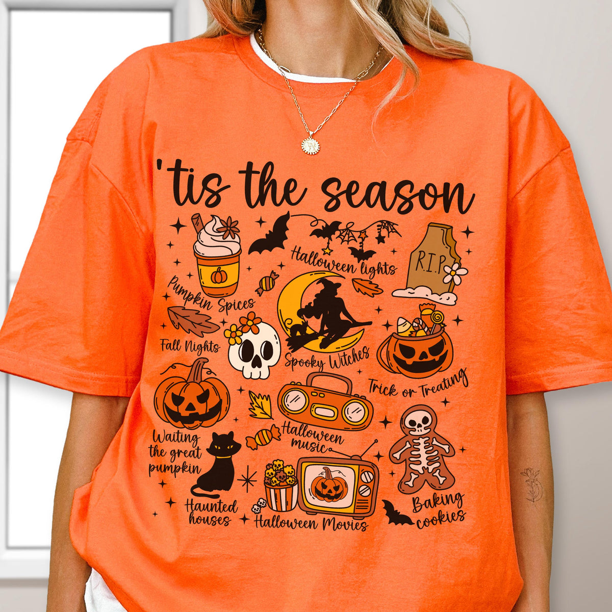 May include: Orange t-shirt with the phrase "'tis the season" and Halloween-themed graphics. The design includes a pumpkin spice latte, bats, a skull, a jack-o'-lantern, and other spooky elements. The shirt is a festive apparel item.
