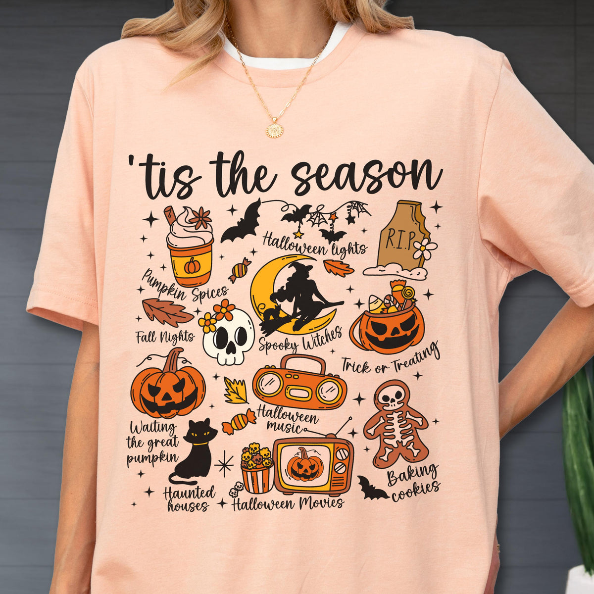 May include: Peach-colored t-shirt with a Halloween-themed graphic. The design includes a witch on a crescent moon, pumpkins, bats, and the text "'tis the season". Other elements include a skull, a cat, and the words "Pumpkin Spices".