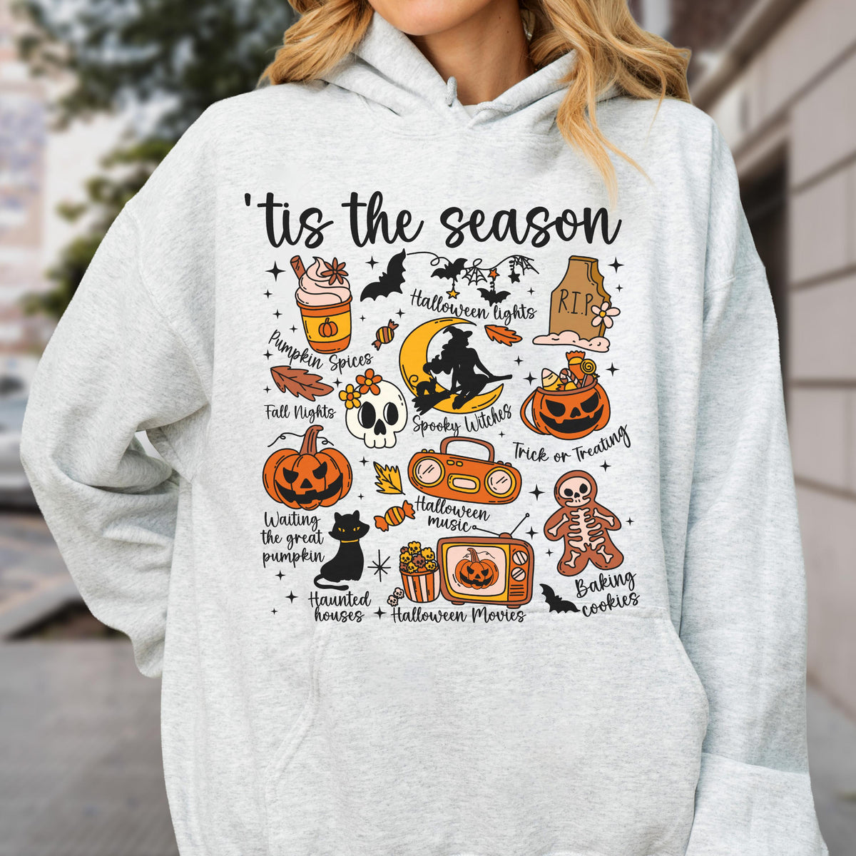May include: A light gray hoodie featuring a colorful Halloween-themed graphic. The design includes pumpkins, bats, a witch, and the phrase "'tis the season." Other elements include "Pumpkin Spices" and "Trick or Treating."