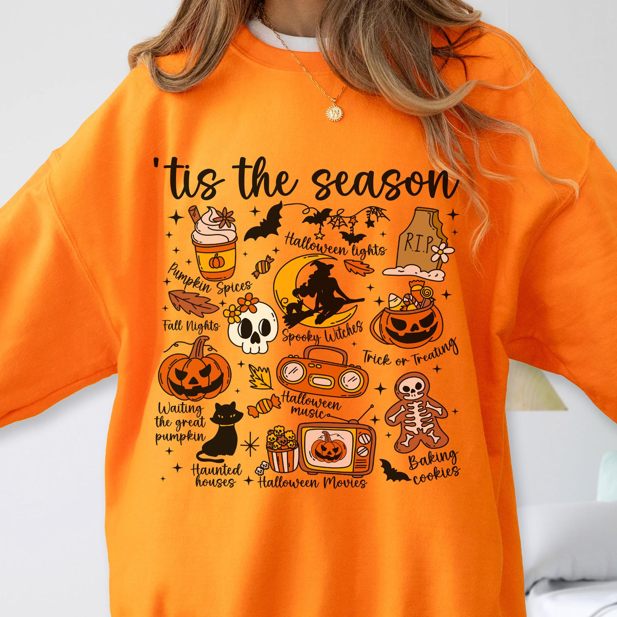 May include: Orange sweatshirt with a Halloween-themed graphic. The design includes a pumpkin spice latte, bats, a skull, a black cat, and the phrase "'tis the season". Other elements include a jack-o'-lantern, a witch, and a tombstone.