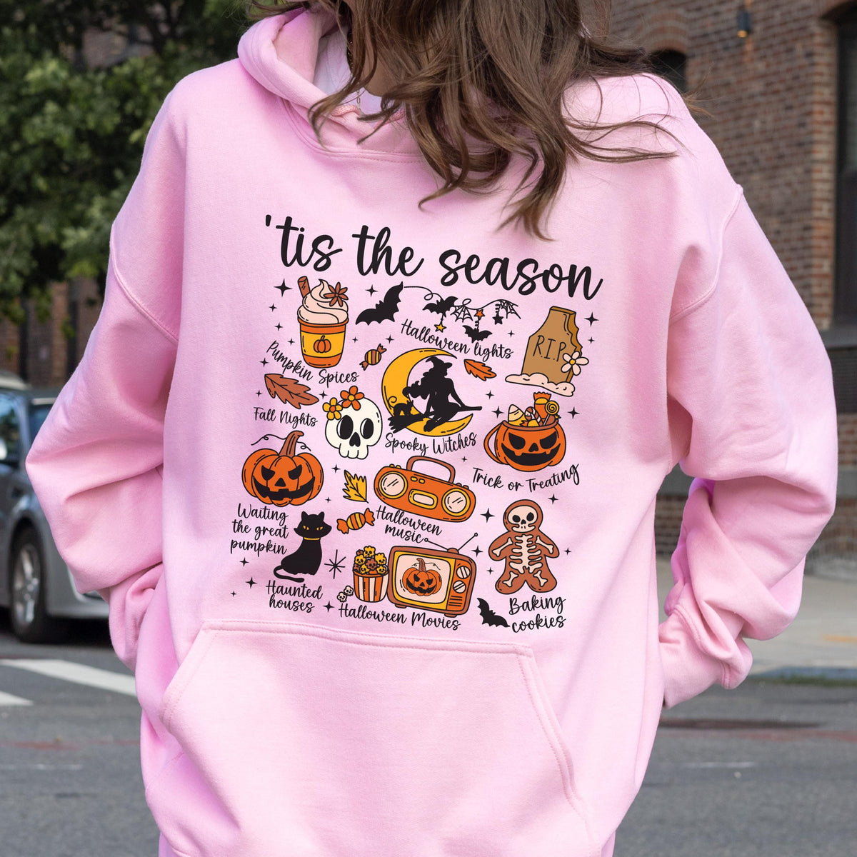 May include: Pink hoodie with a Halloween-themed graphic. The design includes a pumpkin, skull, bats, and the phrase "'tis the season." Other elements include "Pumpkin Spices," "Halloween Lights," and "Trick or Treating."