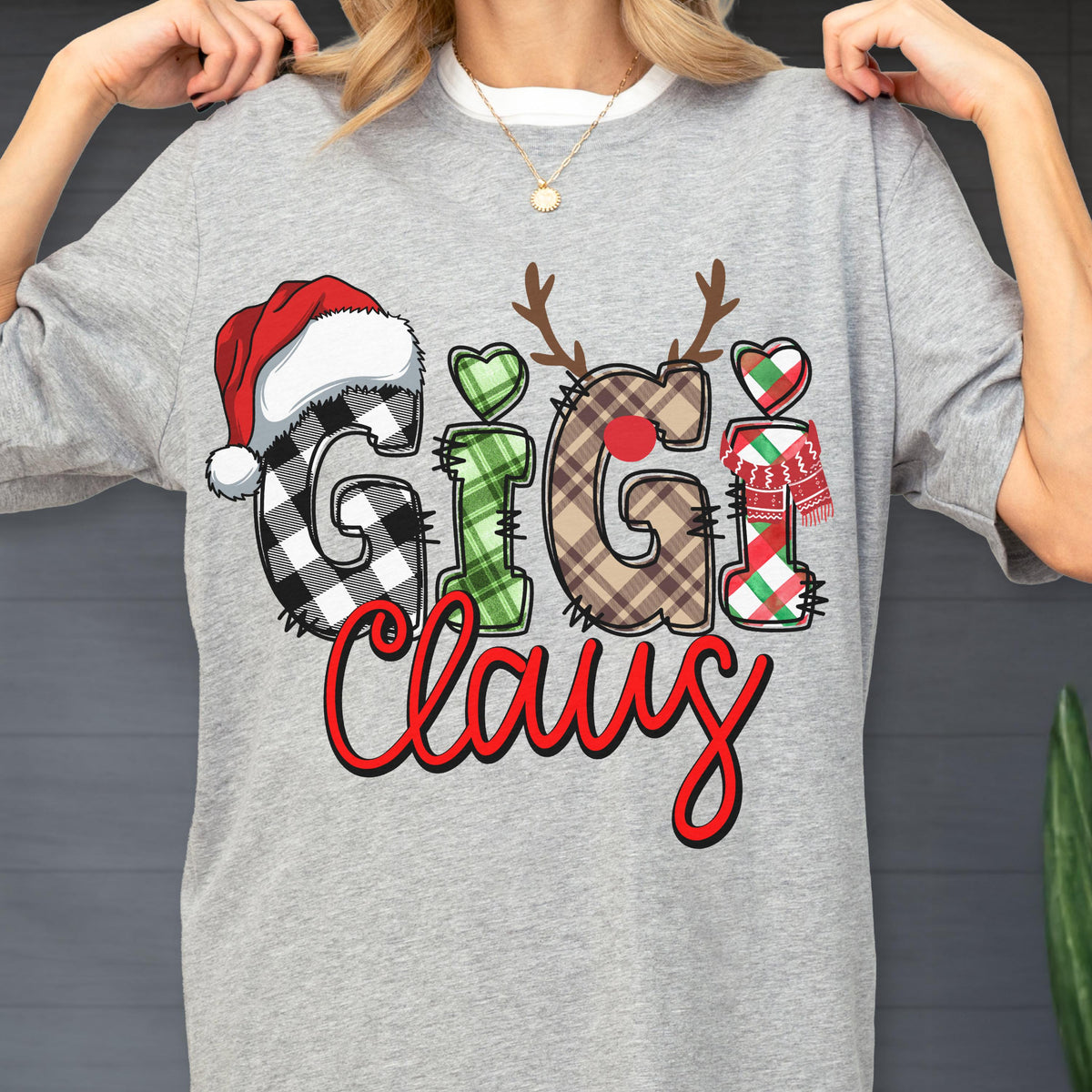 May include: A gray crew neck sweatshirt with the text "Gigi Claus" in a festive design. The letters are decorated with plaid patterns, a Santa hat, reindeer antlers, and a scarf, perfect for the holiday season.