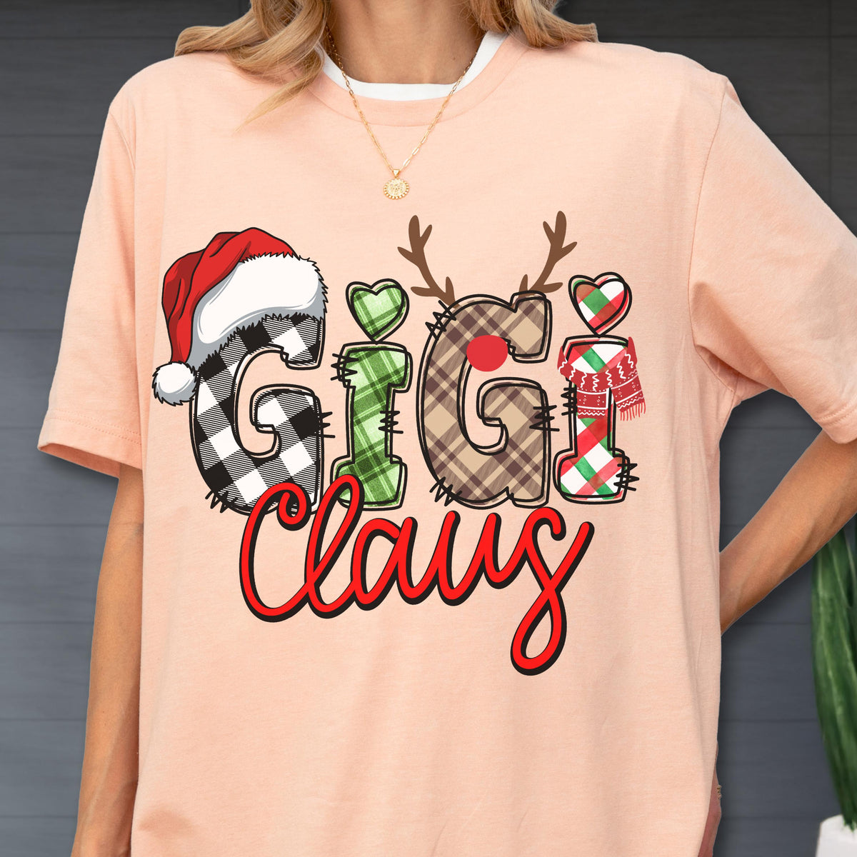 May include: Peach-colored t-shirt with a Christmas-themed graphic. The design features the words "Gigi Claus" in a festive font, with letters decorated as a Santa hat, reindeer antlers, and a scarf. The shirt has a white crew neck.