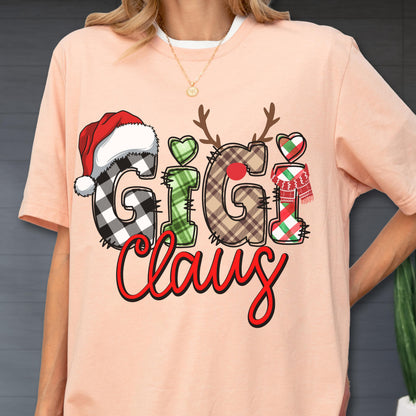 May include: Peach-colored t-shirt with a Christmas-themed graphic. The design features the words "Gigi Claus" in a festive font, with letters decorated as a Santa hat, reindeer antlers, and a scarf. The shirt has a white crew neck.