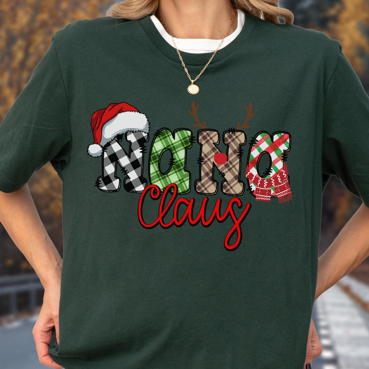 May include: Dark green t-shirt with the text "Nana Claus" in a festive design. The letters are decorated with a Santa hat, reindeer antlers, and plaid patterns in red, green, and black. A gold necklace is visible.