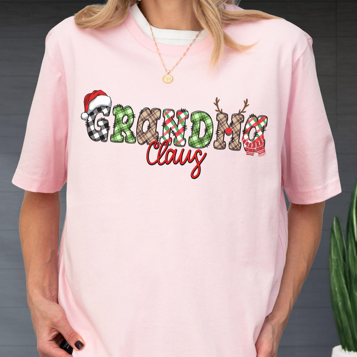 May include: A light pink t-shirt with the words "Grandma Claus" in a festive design. The word "Grandma" is in a plaid pattern, and "Claus" is in a script font. A Santa hat, reindeer antlers, and a scarf are incorporated into the design.