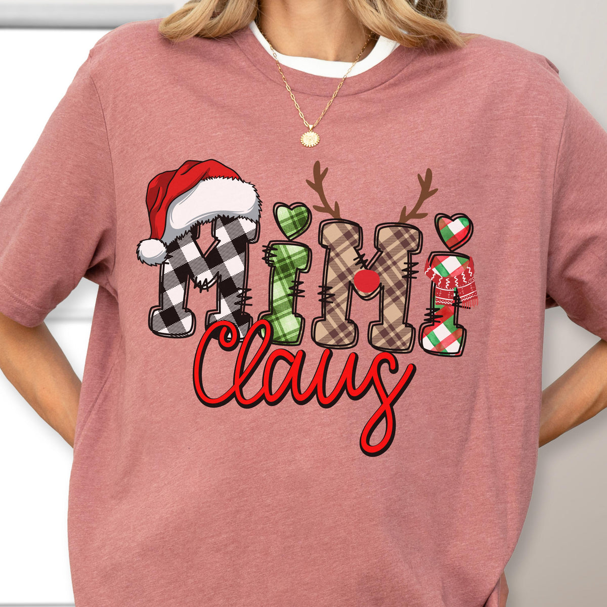 May include: A dusty rose-colored t-shirt with the words "Mini Claus" in a festive design. The letters are decorated with a Santa hat, antlers, hearts, and plaid patterns in red, green, and black. The text is in a playful, handwritten style.