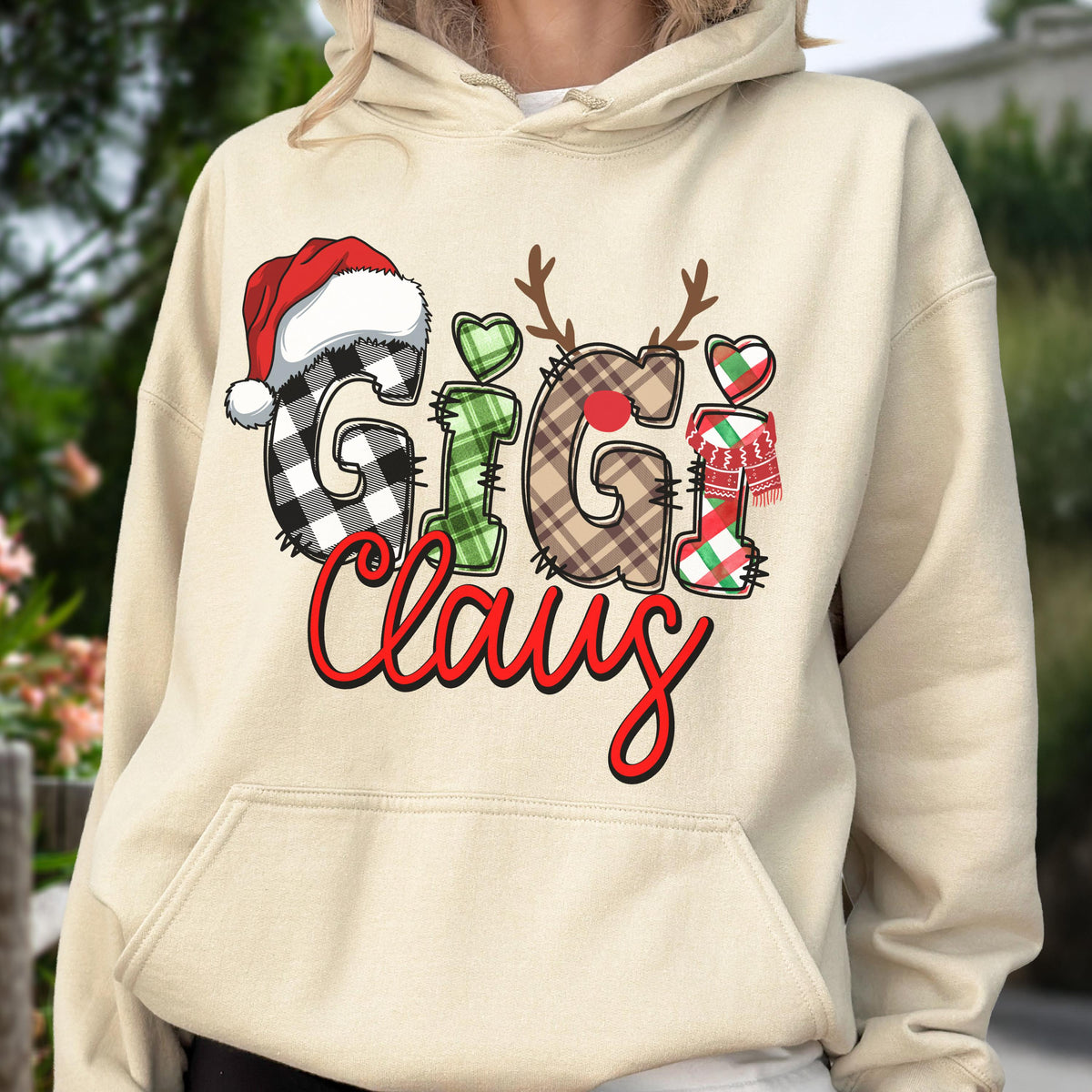 May include: A beige hoodie with a festive "Gigi Claus" design. The design features the words "Gigi Claus" in a mix of fonts and patterns, including a Santa hat, reindeer antlers, and a scarf. The hoodie has a kangaroo pocket.