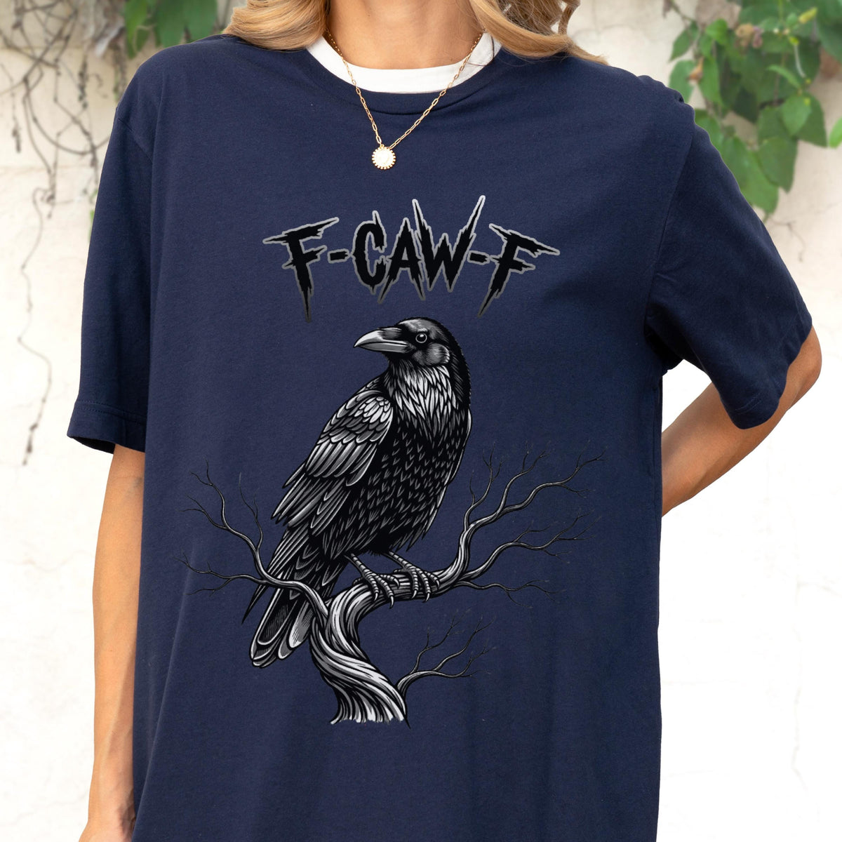 May include: Navy blue t-shirt featuring a detailed illustration of a black raven perched on a tree branch. The text "F-CAW-F" is above the raven. The shirt has a white collar.