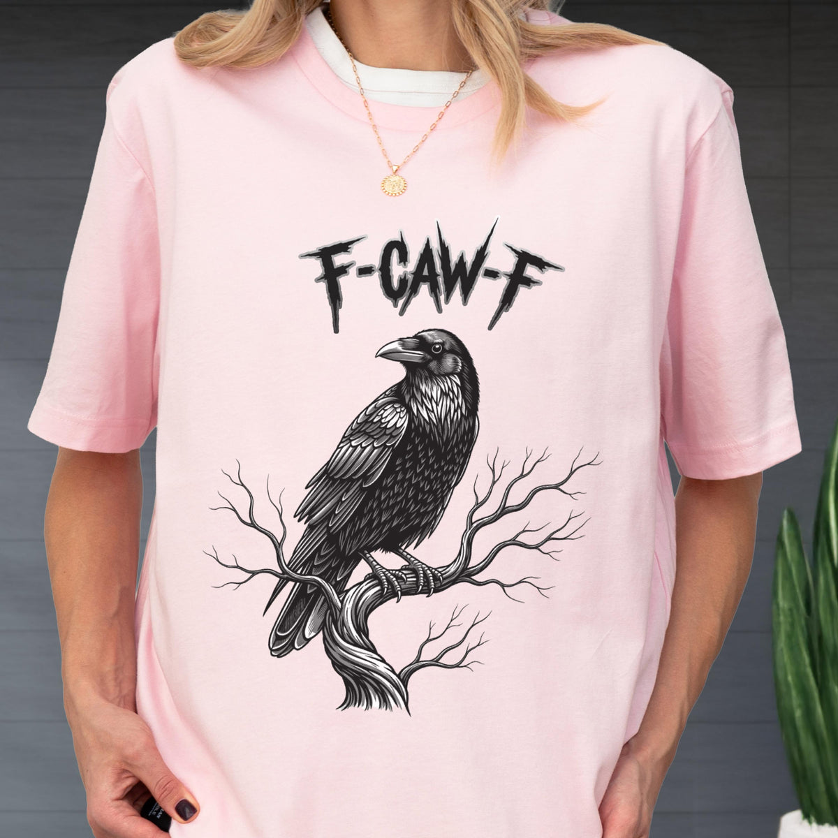 May include: A light pink t-shirt featuring a black and white illustration of a raven perched on a tree branch. The text "F-CAW-F" is above the raven. The shirt has short sleeves and a crew neck.