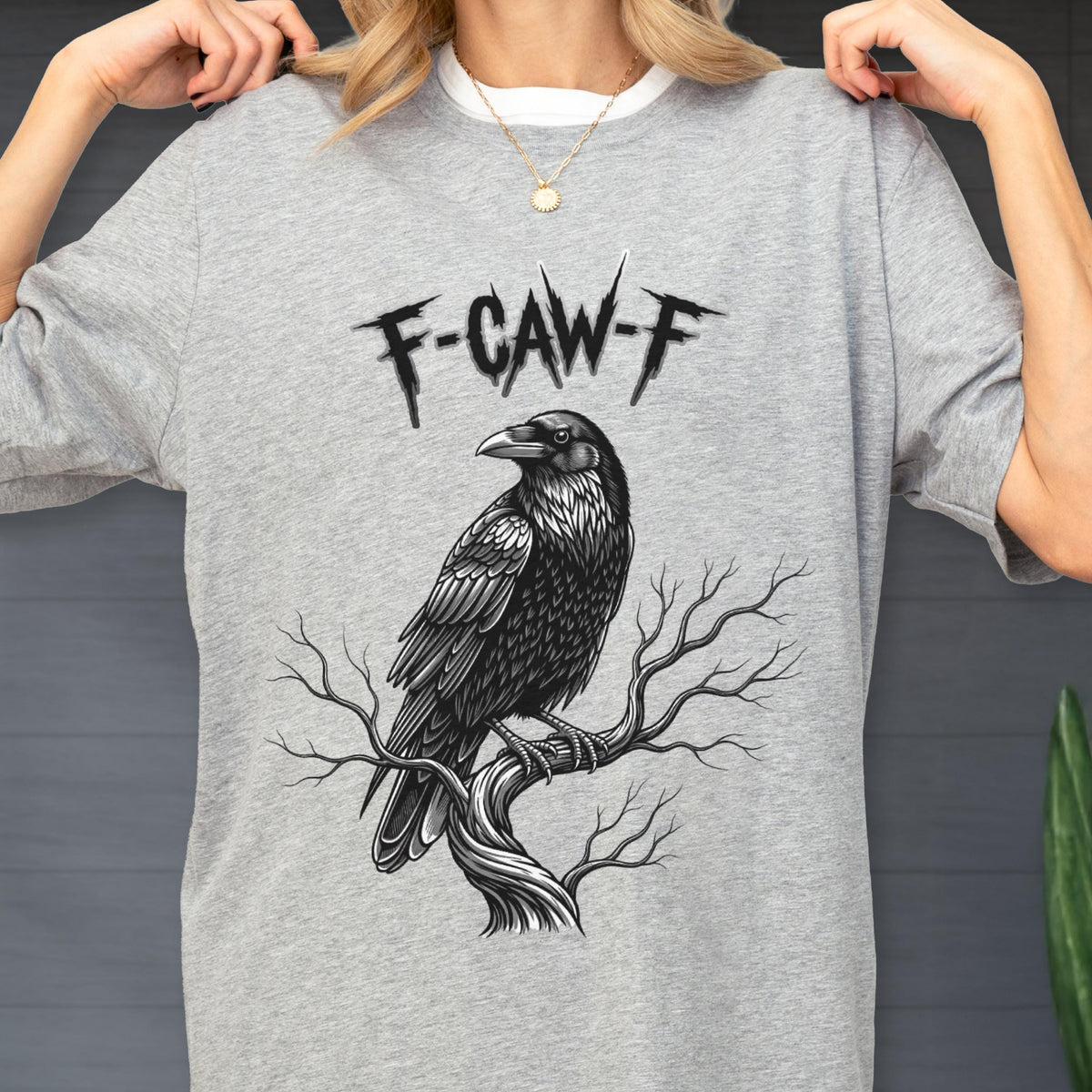May include: A gray t-shirt featuring a detailed black and white illustration of a raven perched on a tree branch. Above the raven is the text "F-CAW-F" in a stylized font. The shirt has a crew neck and short sleeves.