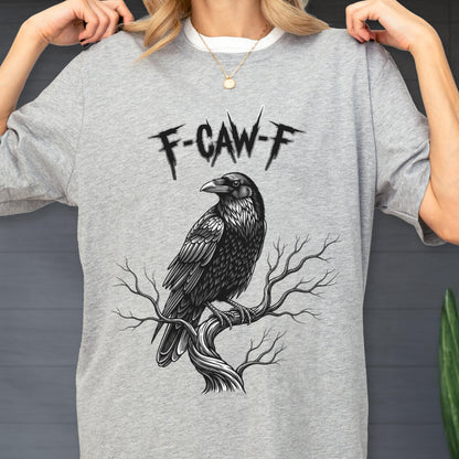 May include: A gray t-shirt featuring a detailed black and white illustration of a raven perched on a tree branch. Above the raven is the text "F-CAW-F" in a stylized font. The shirt has a crew neck and short sleeves.