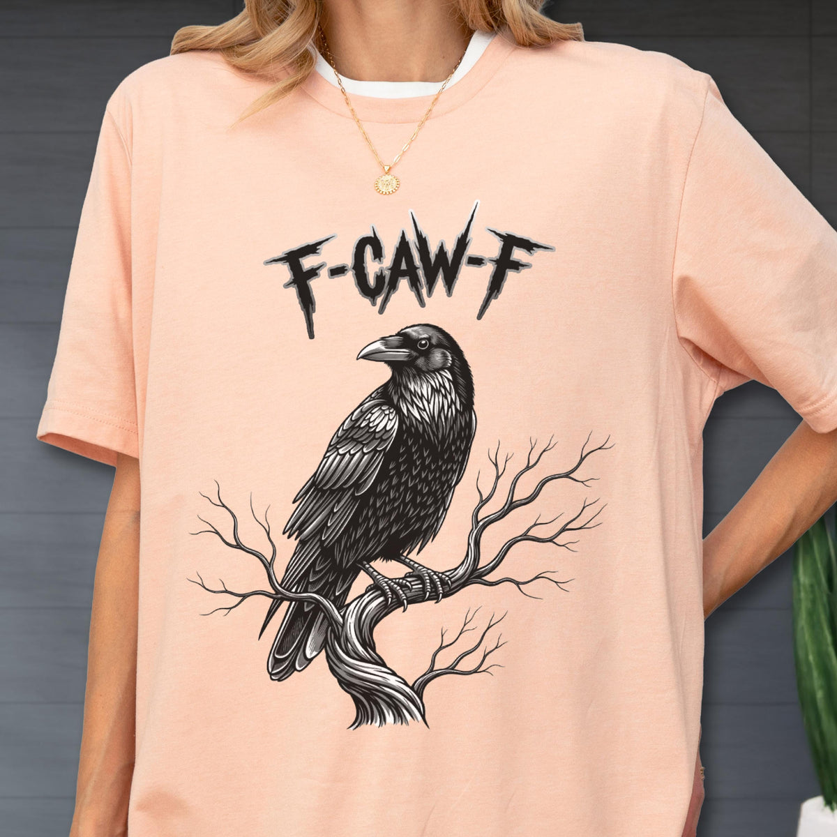 May include: Peach-colored t-shirt featuring a detailed black and white illustration of a raven perched on a tree branch. The text "F-CAW-F" is above the raven. The shirt has a white collar.