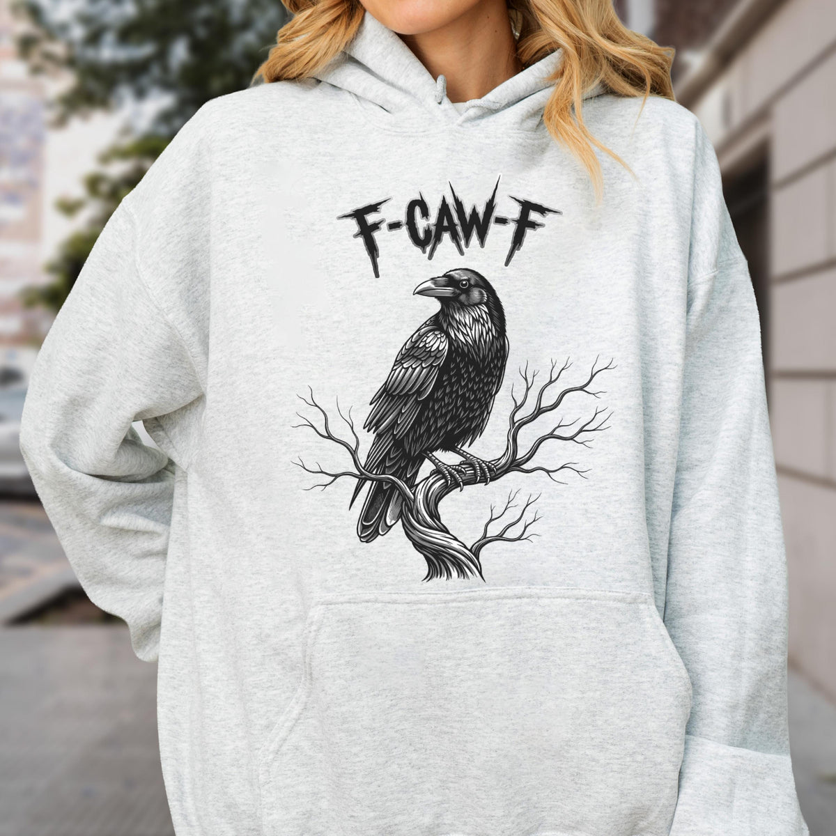 May include: A light gray hooded sweatshirt featuring a black graphic print of a raven perched on a bare tree branch. Above the bird, the text F-CAW-F is printed in a jagged, stylized font. The garment includes a front kangaroo pocket and long sleeves.