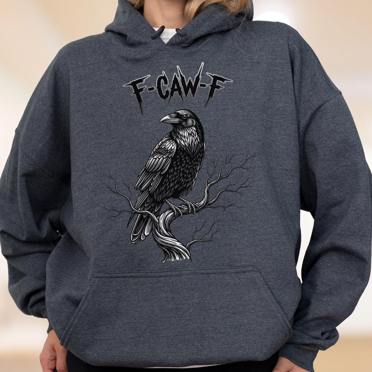 May include: A dark heather gray hooded sweatshirt featuring a graphic illustration of a black raven perched on a bare tree branch. Above the bird, the text F-CAW-F is printed in a stylized, jagged black font. The hoodie includes a front pocket.