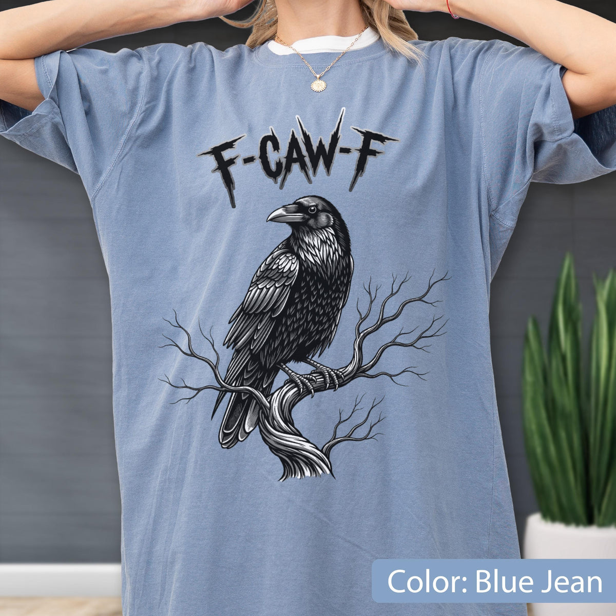 May include: A blue jean colored t-shirt featuring a black and white graphic illustration of a raven perched on a bare tree branch. Above the bird, the text F-CAW-F is printed in a stylized, jagged black font. The shirt has short sleeves.