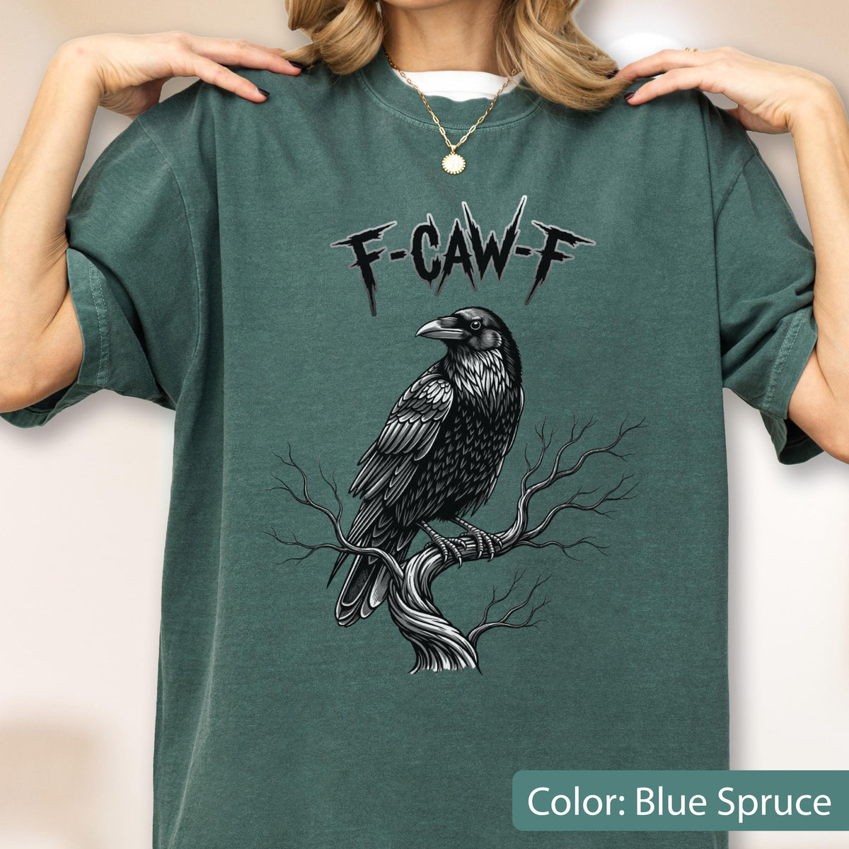 May include: A blue spruce colored t-shirt featuring a graphic print of a black raven perched on a bare tree branch. Above the raven, the text F-CAW-F is printed in a stylized, jagged black font. The shirt has short sleeves and a crew neckline.