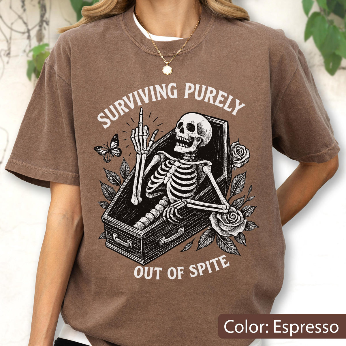 May include: An espresso brown t-shirt featuring a graphic of a skeleton inside a coffin, giving the middle finger. The text reads "SURVIVING PURELY OUT OF SPITE" in white lettering, surrounded by illustrations of a butterfly and two roses.