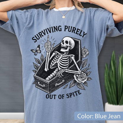 May include: A blue jean colored t-shirt featuring a black graphic print of a skeleton inside a coffin, giving the middle finger. The text reads SURVIVING PURELY OUT OF SPITE. The design includes illustrations of a butterfly and two roses.